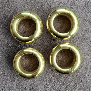 Puede incluir: Cuatro anillos de cortina de metal de color dorado. Los anillos son redondos, con una superficie lisa y un agujero en el centro.
