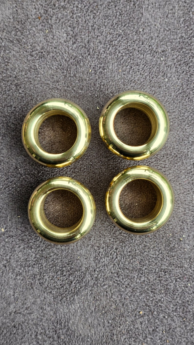 Puede incluir: Cuatro anillos de cortina de metal de color dorado. Los anillos son redondos, con una superficie lisa y un agujero en el centro.