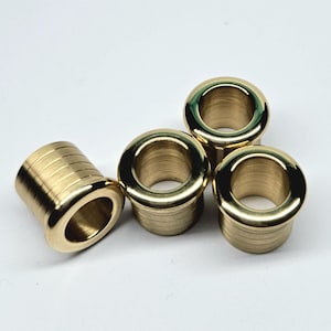 Può includere: Quattro occhielli in ottone lucido disposti su una superficie bianca. Ogni occhiello ha un corpo cilindrico con un bordo svasato e arrotondato e un foro centrale. Gli occhielli sono progettati per rinforzare i fori in tessuto o altri materiali.