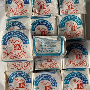 May include: A stack of 12 white bars of soap with blue and red lettering. The soap is labeled "AL-JAMAL SOAP OLIVE OIL SOAP SHOWER SOAP" and features a camel logo. The text is in Arabic.
