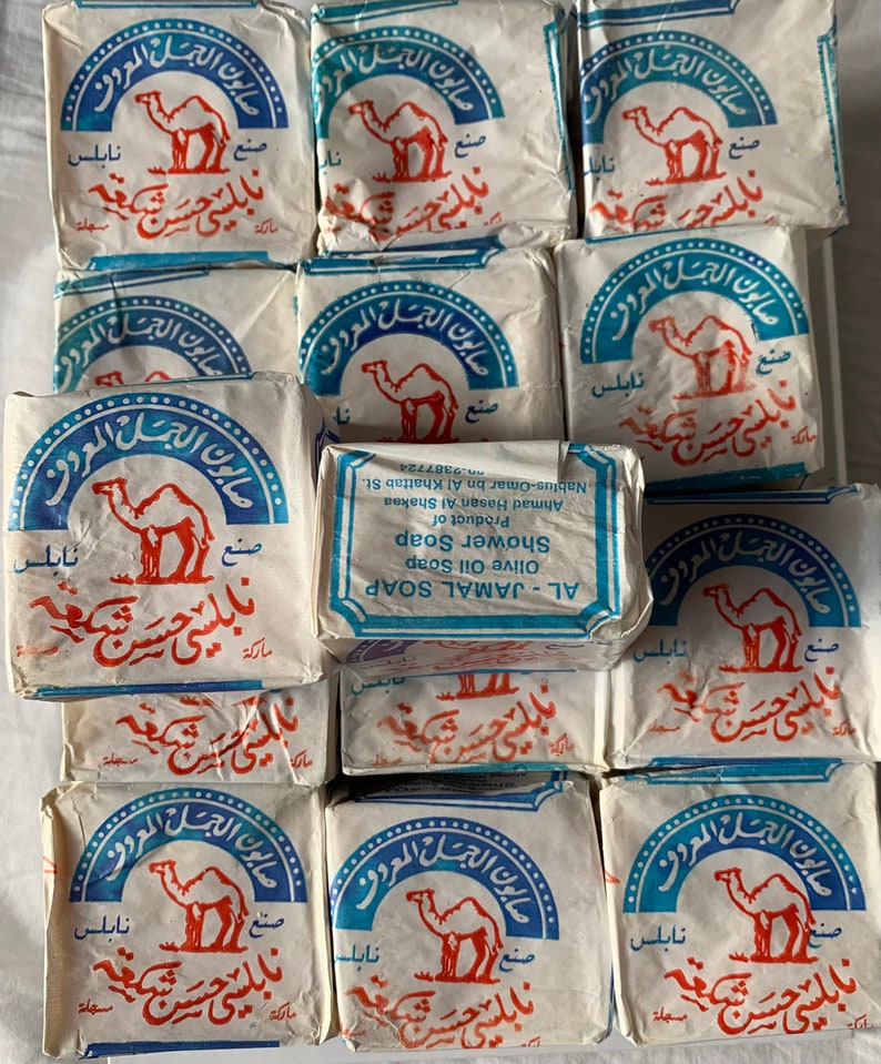 May include: A stack of 12 white bars of soap with blue and red lettering. The soap is labeled "AL-JAMAL SOAP OLIVE OIL SOAP SHOWER SOAP" and features a camel logo. The text is in Arabic.
