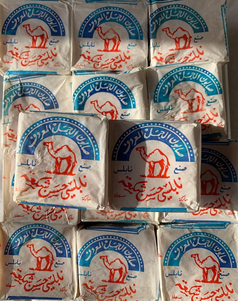 May include: A stack of white rectangular soap bars with blue and red text and a camel graphic. The text reads "صابون الجمل المعروف" which translates to "The Famous Camel Soap".