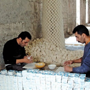 May include: Two people are working in a workshop, making and packaging bars of soap. The soap is stacked in piles and arranged in rows. The soap is white and has a rough texture. The people are wearing casual clothing.