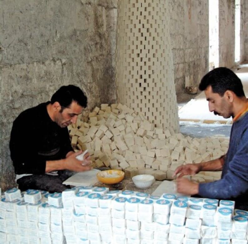 May include: Two people are working in a workshop, making and packaging bars of soap. The soap is stacked in piles and arranged in rows. The soap is white and has a rough texture. The people are wearing casual clothing.