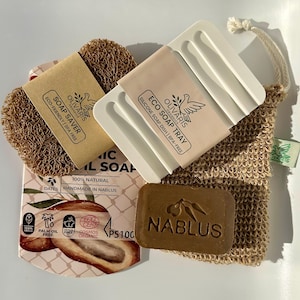 Puede incluir: Un surtido de productos de jabón natural, que incluye una pastilla de jabón marrón con la palabra "Nablus" en relieve, un jabonera de silicona blanca, un salvador de jabón marrón y una bolsa de malla. El embalaje incluye el texto "Eco-Friendly | BPA Free".