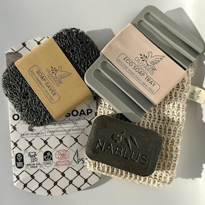 May include: A selection of natural soap products, including a dark grey soap saver, a grey silicone soap tray, a bar of soap with the word "Nablus" embossed, and a mesh soap bag. The packaging features the "Olivaris Organics" logo.