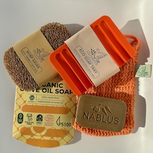 Puede incluir: Un surtido de productos de baño ecológicos, que incluyen un salvador de jabón marrón, un jabonera de silicona naranja, una pastilla de jabón de aceite de oliva y una bolsa de malla naranja. La jabonera y el salvador de jabón tienen etiquetas con la marca "Olivaris Organic".