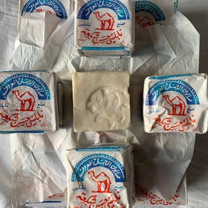 May include: Six bars of camel soap wrapped in white paper. The soap is white and has a camel printed on the wrapper. The wrapper is blue and white with Arabic text.