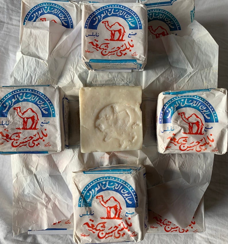 May include: Six bars of camel soap wrapped in white paper. The soap is white and has a camel printed on the wrapper. The wrapper is blue and white with Arabic text.