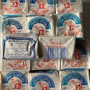 May include: A close-up of a stack of white soap bars with blue and red printed labels. The labels feature a camel and the text "صابون الجمل المعروف" in Arabic. One bar has a white label with the text "PS 101" and a barcode. The text "Regestration No رقم التشغيلة" and "للاستحمام" is also visible on the label.