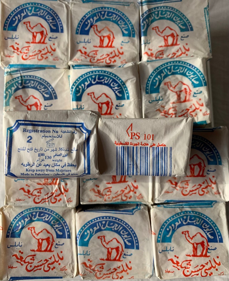 May include: A close-up of a stack of white soap bars with blue and red printed labels. The labels feature a camel and the text "صابون الجمل المعروف" in Arabic. One bar has a white label with the text "PS 101" and a barcode. The text "Regestration No رقم التشغيلة" and "للاستحمام" is also visible on the label.
