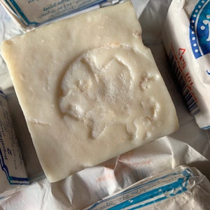 May include: A white bar of soap with a raised design of a camel on the top. The soap is wrapped in blue and white paper with Arabic text that reads "صابون الجمل" which translates to "Camel Soap".