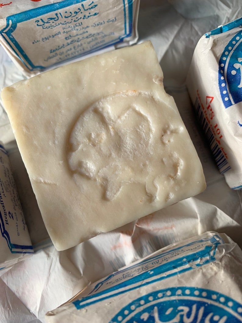 May include: A white bar of soap with a raised design of a camel on the top. The soap is wrapped in blue and white paper with Arabic text that reads "صابون الجمل" which translates to "Camel Soap".