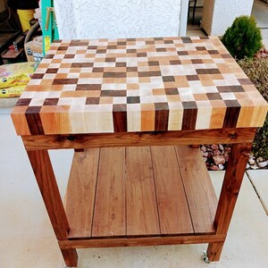May include: A wooden butcher block table with a checkered top made of different wood types. The table has a lower shelf and is on wheels.