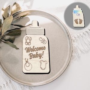 Könnte beinhalten: Ein hölzernes Willkommens-Schild in Form einer Babyflasche mit dem Text "Willkommen Baby!" und kleinen gravierten Bildern von einem Schnuller, einer Rassel und einem Strampler.