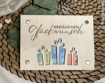May include: A wooden gift card holder with the German phrase "HERZLICHEN Glückwunsch" cut out. The design includes cut-out gift boxes, each revealing colorful Euro banknotes. Two small wooden hearts and greenery are also visible.