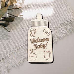 May include: Wooden baby bottle shaped sign with the text "Welcome Baby!" and engraved baby icons.