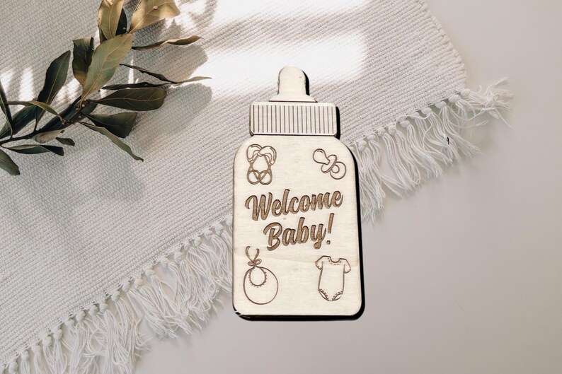 May include: Wooden baby bottle shaped sign with the text "Welcome Baby!" and engraved baby icons.