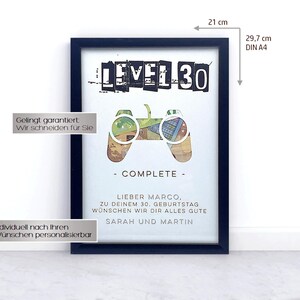 Op de afbeelding: Een zwart ingelijste poster met een witte achtergrond. De poster heeft een zwart-witte afbeelding van een videogamecontroller met de tekst "LEVEL 30" erboven. De tekst "COMPLETE" staat onder de controller. De poster bevat ook de tekst "Lieber Marco, zu deinem 30. Geburtstag w&uuml;nschen wir dir alles gute Sarah und Martin".