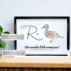 May include: A framed print featuring the text ENDLICH R- and a geometric duck illustration filled with multicolored patterns. Below the duck, the text reads Wir werden Dich vermissen! ZUM ABSIED ALLES GUTE. The frame is silver and rectangular.