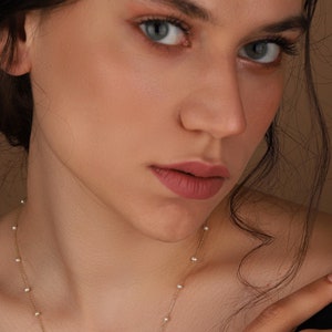 May include: A woman with dark hair and brown eyes is wearing a delicate gold necklace with small white pearls. The necklace is visible against her bare shoulders and neck.