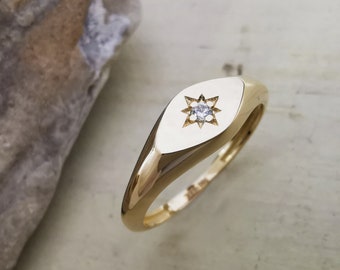Puede incluir: Un anillo de sello dorado con un diseño de estrella. El anillo presenta una banda de oro pulido y una cara ovalada y plana. La estrella está engastada con una pequeña gema transparente. El anillo está sobre una superficie clara.