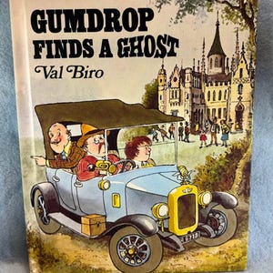May include: A vintage book cover titled "Gumdrop Finds a Ghost" by Val Biro. The illustration depicts a light blue car with three passengers driving past a castle. The background features a detailed castle and figures.
