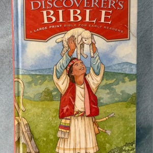 May include: A hardcover "Discoverer's Bible" for early readers. The cover features an illustration of a person holding a lamb, with a red vest and white tunic. The title is in a red and white banner, with the text "New International Reader's Version".
