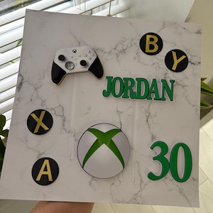 May include: A white and green Xbox controller cake topper with the words "By Jordan 30" on a marble background.
