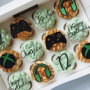 May include: A box of cupcakes decorated with green and black frosting and sprinkles. The cupcakes have various designs, including a video game controller, headphones, and the words "Happy Birthday" and "Charlie".