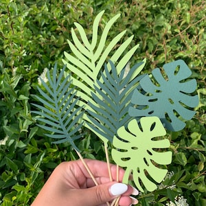 May include: A selection of paper tropical leaves in various shades of green, including a monstera leaf and fern-like designs, attached to wooden skewers. The leaves are held against a backdrop of green foliage.