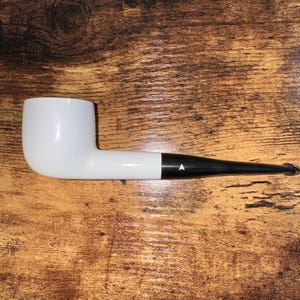 May include: A white tobacco pipe with a black stem and a small white triangle detail. The pipe is set against a wooden background. The pipe is a classic smoking accessory.