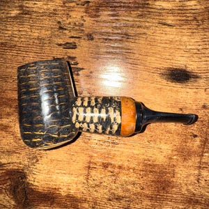 May include: A tobacco pipe with a dark, textured bowl and a patterned stem. The pipe features a black mouthpiece and an orange accent. The pipe is made of wood and is designed for smoking tobacco.