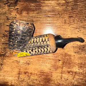 May include: A corn cob pipe with a black stem and a textured bowl. The bowl has a dark, patterned design. The pipe rests on a wooden surface. A yellow sticker with text is visible on the pipe.