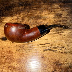 May include: A brown tobacco pipe with a smooth, polished finish and a curved black stem. The bowl features a rounded shape with a subtle rim detail. The side of the bowl is stamped with the text E. WILKE N.Y.C. in a clear, legible font.