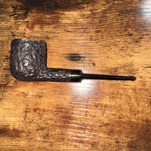 May include: A dark brown tobacco pipe with a textured bowl and a long, black stem. The pipe rests on a wooden surface with visible grain patterns. The pipe's bowl has a rough, bark-like texture.