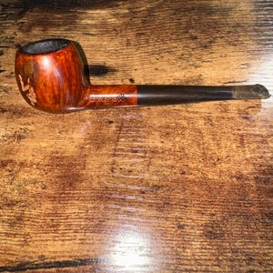 May include: A wooden tobacco pipe with a curved bowl and a long, dark stem. The bowl is a warm brown color with a lighter, reddish-brown area. The stem is dark brown with a silver tip. The pipe is resting on a wooden surface.