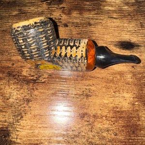 May include: A corn cob pipe with a black stem and a textured, patterned bowl. The bowl is primarily black and yellow, with a cylindrical shape. The pipe rests on a wooden surface.