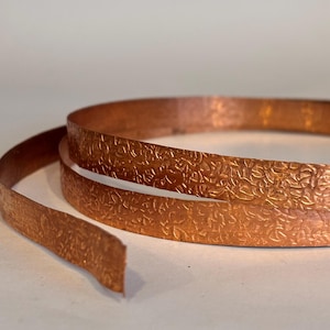 May include: A coiled copper strip featuring a textured floral pattern. The copper has a warm, reddish-brown hue and a slightly reflective surface. The strip is unrolled, revealing its length and the intricate details of the embossed design.