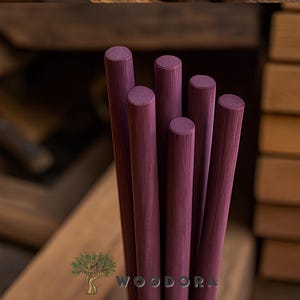 May include: A close-up of six cylindrical wooden dowels in a deep burgundy color. The dowels are arranged closely together, with their rounded ends visible. The wood grain is subtly visible. The logo "WOODORA" is at the bottom.