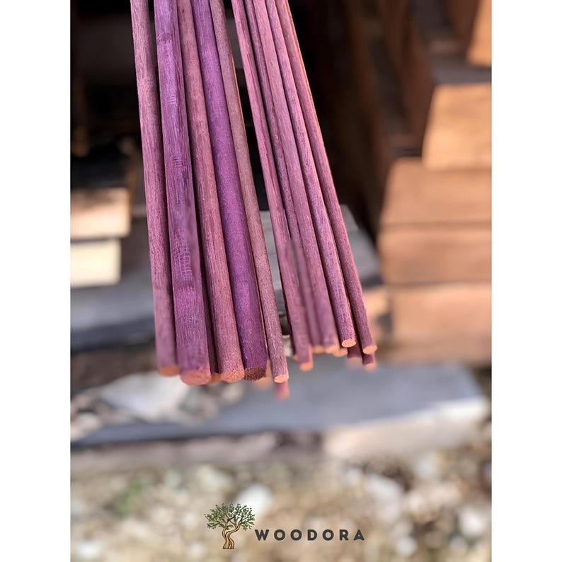 May include: A bundle of purple wooden dowels, each with a rounded end. The dowels are arranged in a fan shape, with the ends of the dowels visible. The wood has a natural grain and a rich, saturated color. The logo "WOODORA" is visible at the bottom.