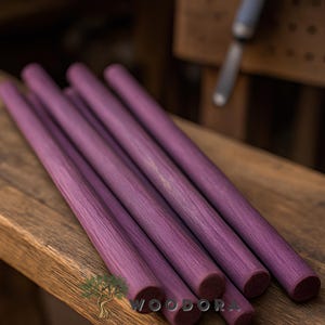 May include: A collection of six purple wooden dowels, arranged diagonally on a wooden surface. The dowels have a textured grain and rounded ends. The word "WOODORA" is visible in the bottom right corner.