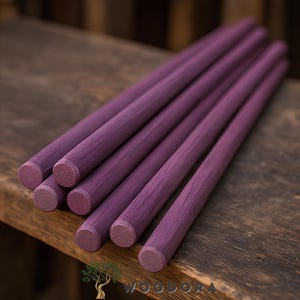 May include: A collection of seven purple wooden dowels, each with a rounded end. The dowels are arranged diagonally on a dark wooden surface. The wood grain is visible, and the color is a deep, saturated purple.