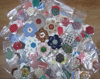 May include: A collection of colorful, fabric hexagon shapes in clear plastic bags. The shapes are various sizes and patterns, including floral, geometric, and novelty designs. The background is a wooden surface.