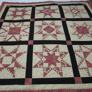 May include: A handmade quilt featuring a patchwork design with red and cream-colored star patterns. The stars are set against a cream background with a paisley print, separated by black borders. The quilt has a red border.