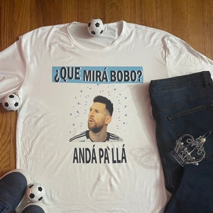 May include: White t-shirt with a graphic of Lionel Messi in a soccer jersey. The text on the shirt reads "¿QUE MIRÁ BOBO? ANDÁ PA' LLÁ".