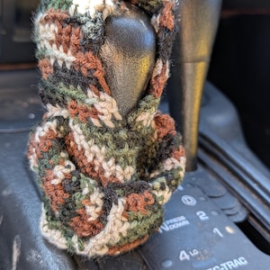 Könnte beinhalten: Ein kleiner, gehäkelter Kapuzenpullover-Überzug für einen Schaltknauf mit einem tarnfarbenen Muster in Waldgrün, Dunkelbraun, Schwarz und Creme. Das Kleidungsstück ist aus Garn gefertigt und verfügt über eine kleine Kapuze und lange Ärmel.