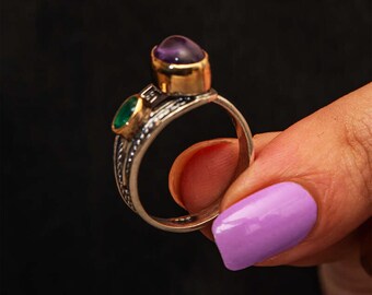 May include: A silver ring with gold accents, featuring a large oval amethyst and a smaller emerald gemstone. The ring is held against a dark background, showcasing its intricate design and colorful gemstones.