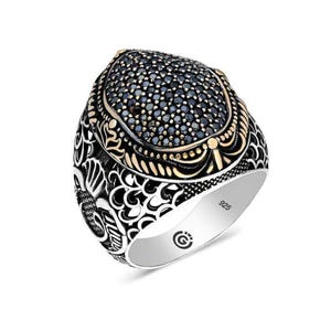 Puede incluir: Un anillo de plata con una piedra negra engastada en un diseño de filigrana dorado. El anillo está marcado con "925" en la banda interior.