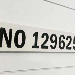 May include: A rectangular white sign with the text "NO 1296259" in bold black letters. The sign is mounted on a white wall with horizontal paneling. The sign has rounded corners and small black dots at each corner.